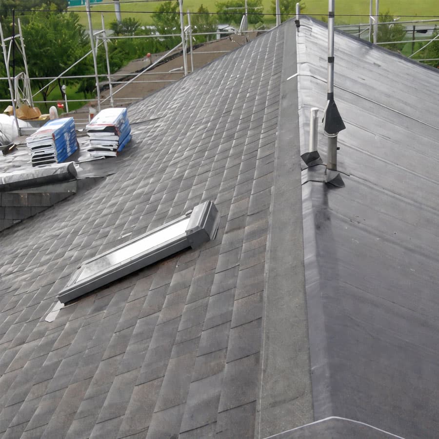Vista ravvicinata di un tetto in fase di ristrutturazione con nuove tegole scure, un lucernario moderno installato e impalcature di sicurezza sullo sfondo.