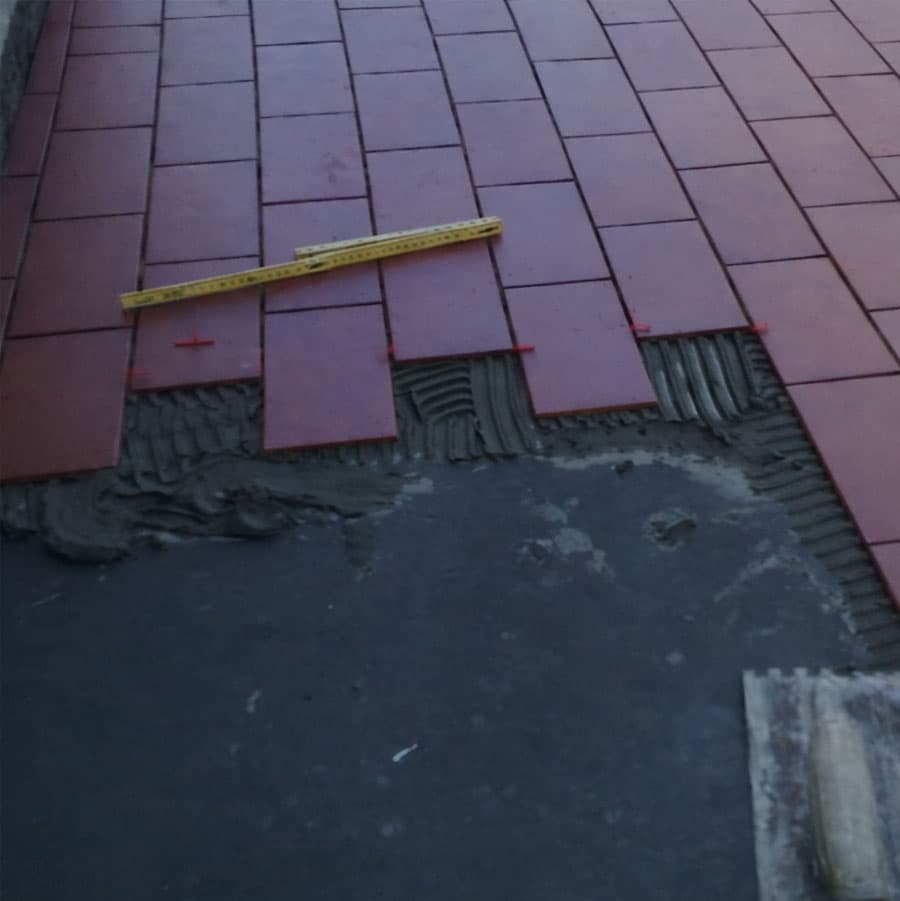 Fase di posa di piastrelle in klinker o gres su uno strato di guaina impermeabilizzante per terrazzi, con dettaglio del collante steso e strumenti di misurazione professionale.